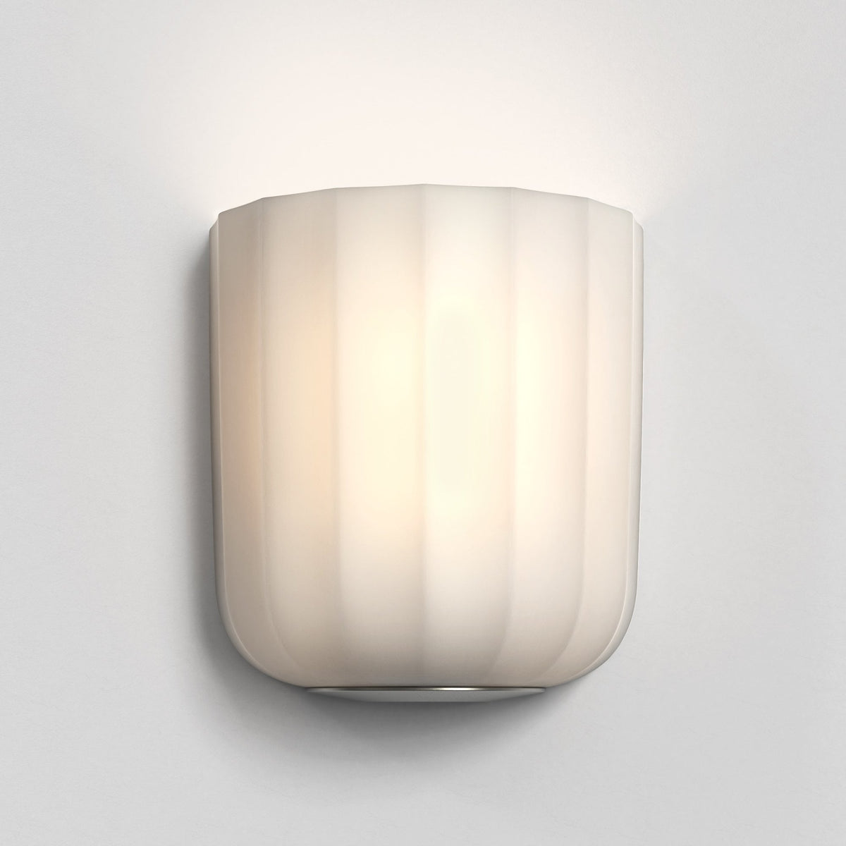 White fluted wall sconce with a warm glow mounted on a light gray wall