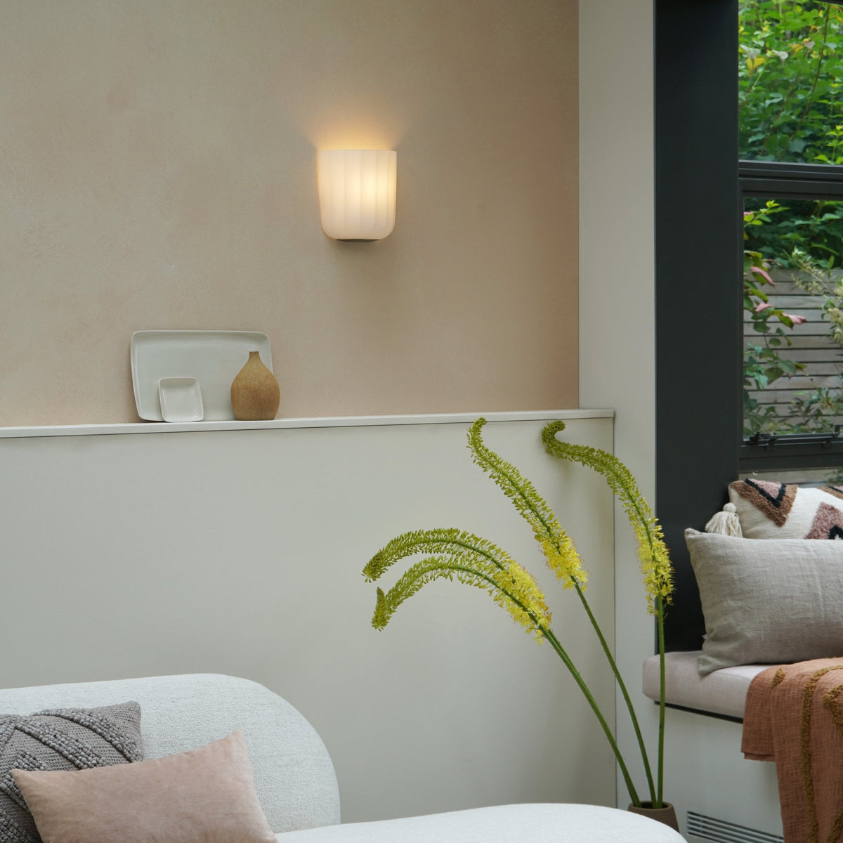 Wall sconce with a ribbed white shade illuminating a beige wall above a white shelf with decor