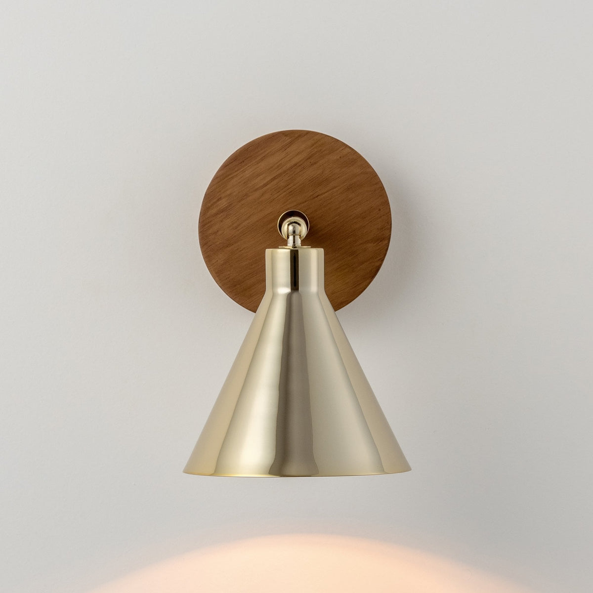 A modern wall lamp with a brass conical shade and a round wooden base, casting light.