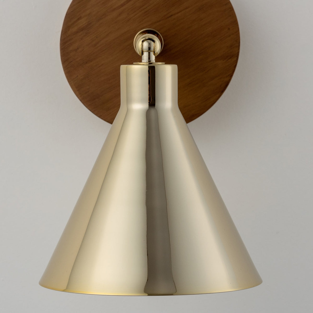 A front view of a modern wall lamp with a brass conical shade and a round wooden base.