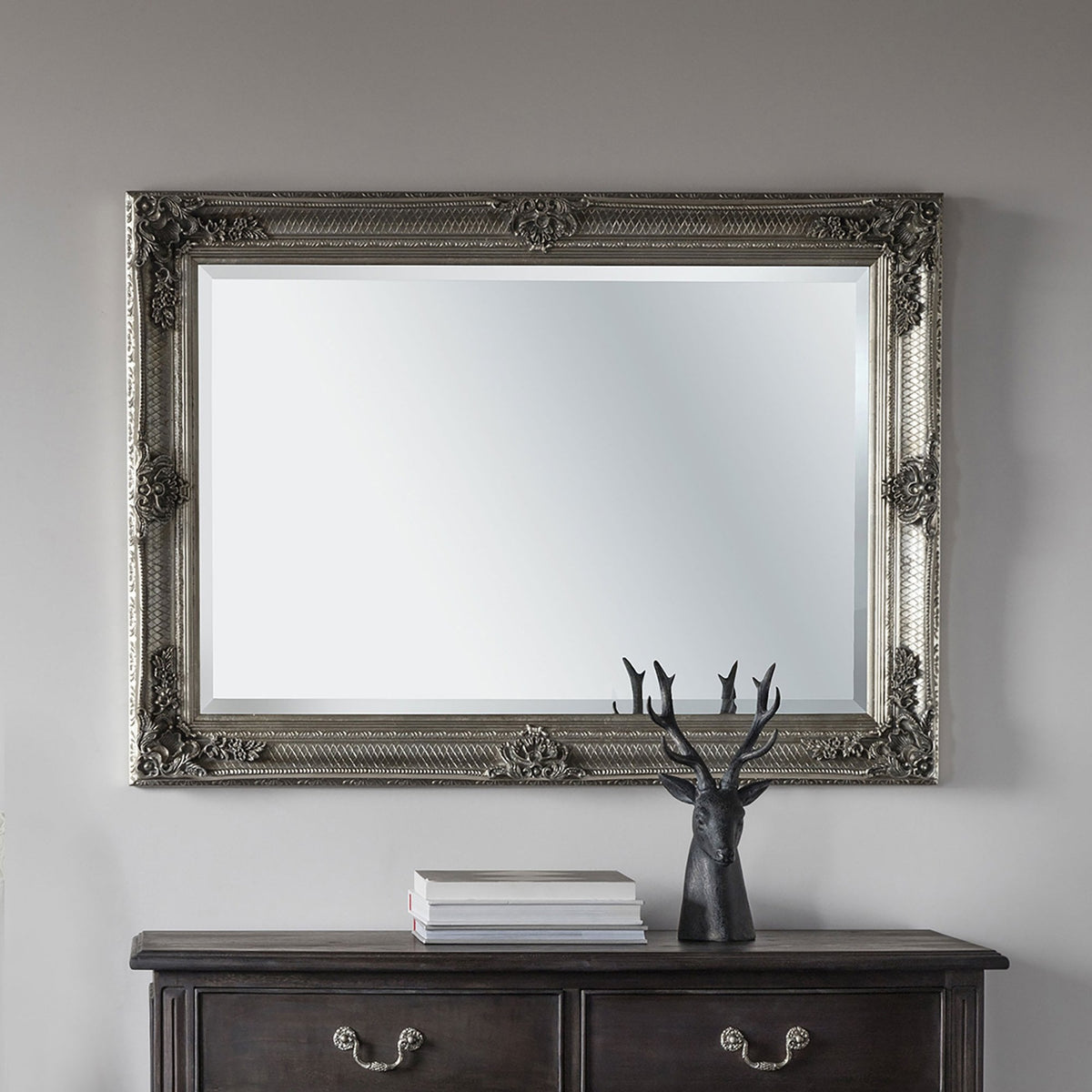 A horizontal ornate silver mirror hangs over a dark wooden chest of drawers with a stag ornament.