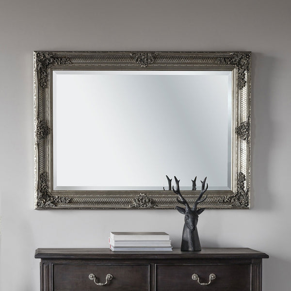 A horizontal ornate silver mirror hangs over a dark wooden chest of drawers with a stag ornament.