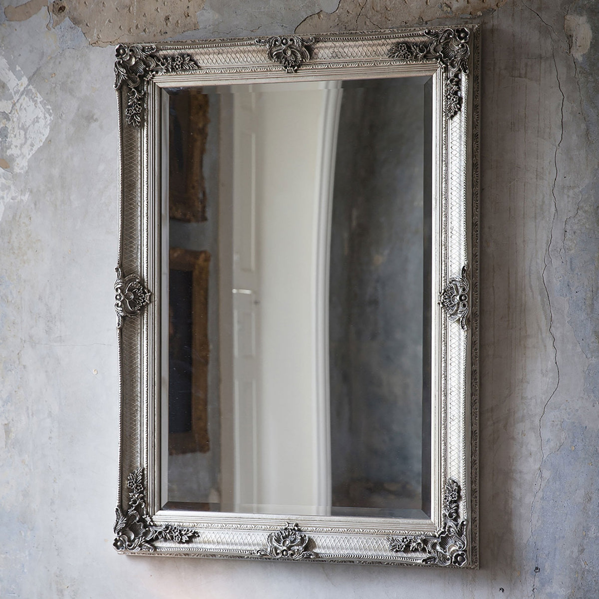 A tall, ornate silver mirror leans against a textured, light grey wall.