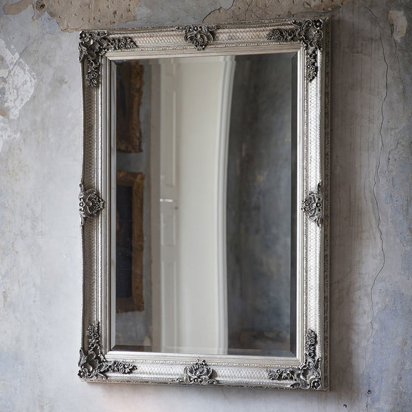 A tall, ornate silver mirror leans against a textured, light grey wall.
