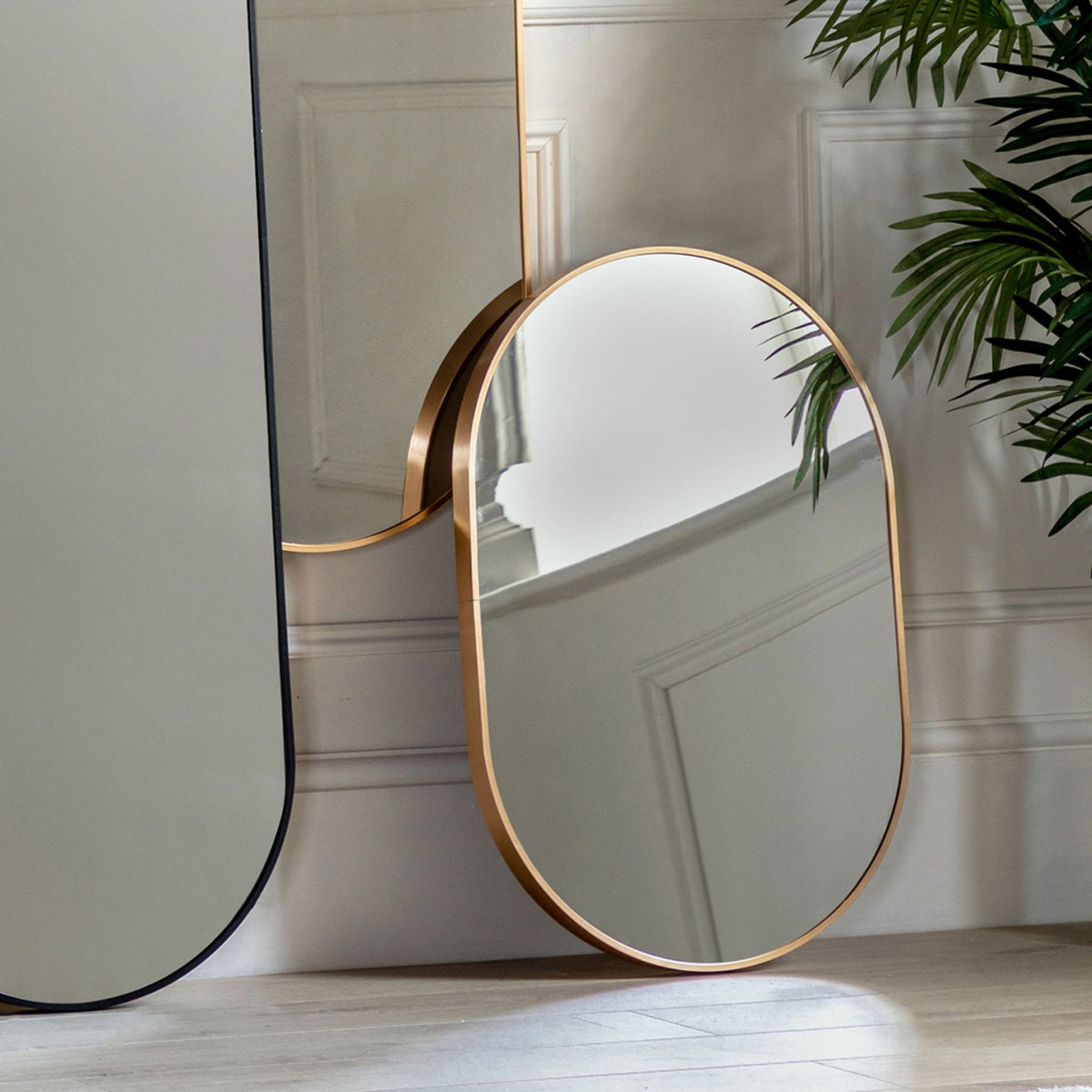 A close-up of an oval gold-framed mirror leaning against a white panelled wall.