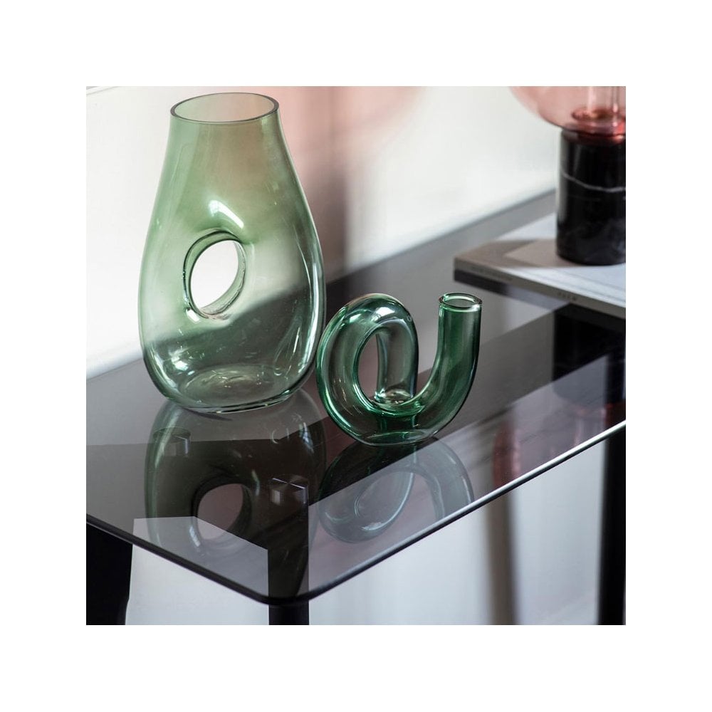 Two green glass vases, one with a hole, the other S-shaped, on a glass table top.