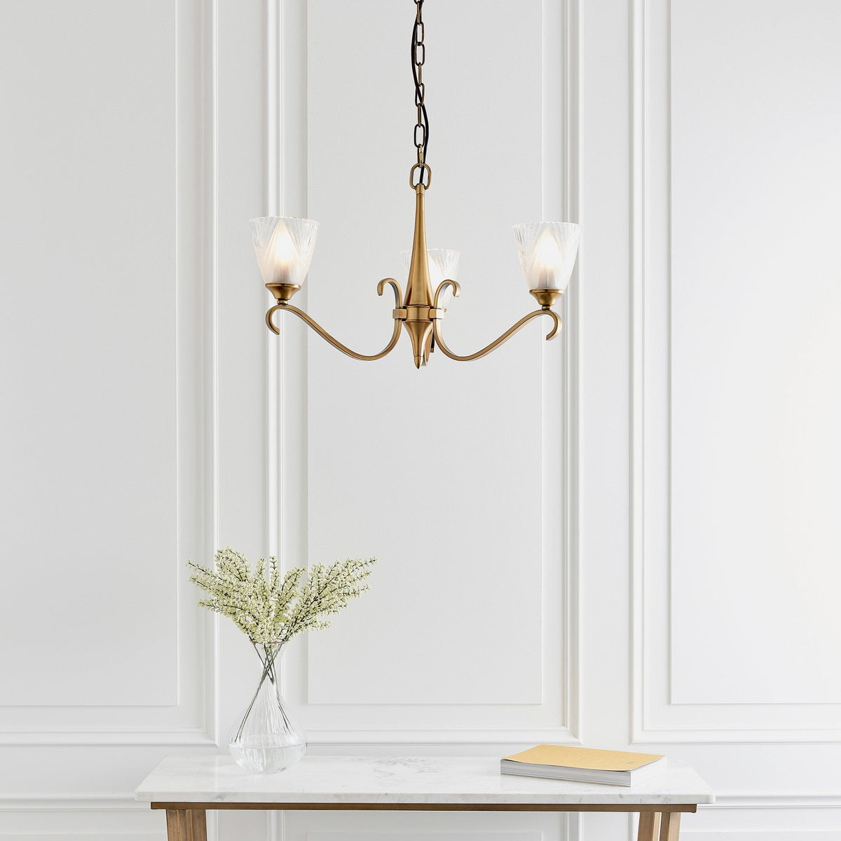 A three-light brass chandelier with frosted glass shades hangs above a white console table.