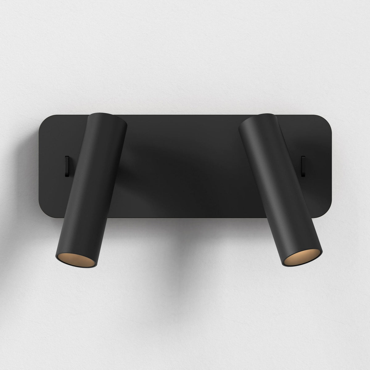 Black rectangular wall light with two cylindrical spotlights angled downwards, mounted on a white wall