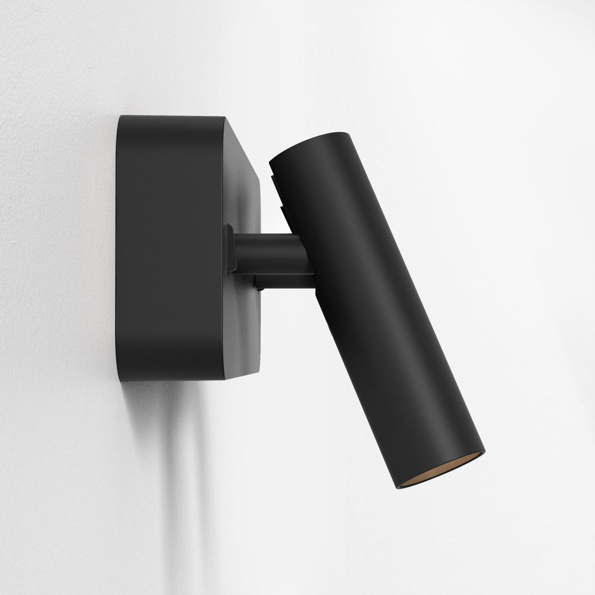 Black wall-mounted spotlight with a cylindrical head angled downwards, against a white textured wall