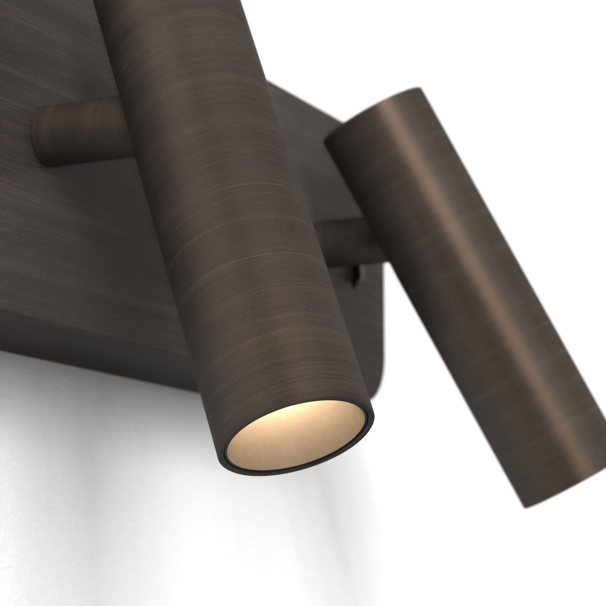 Two dark bronze cylindrical wall lights with one illuminated, casting a warm glow on a white wall