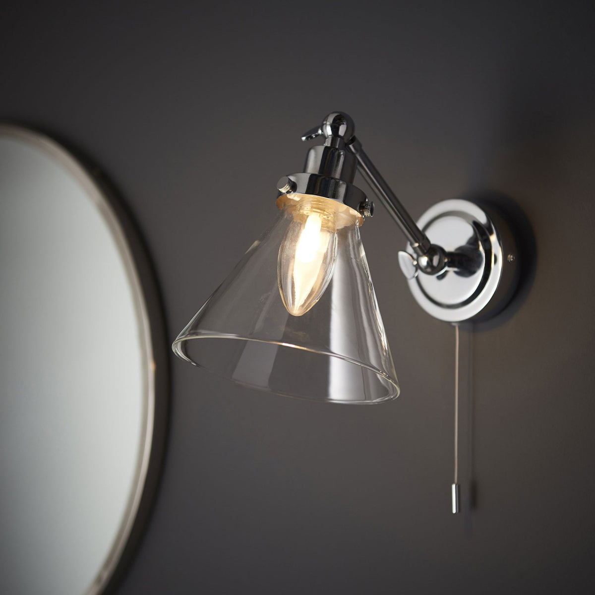 Chrome wall light with clear glass cone shade and pull cord switch, illuminating a dark grey wall next to a round mirror.