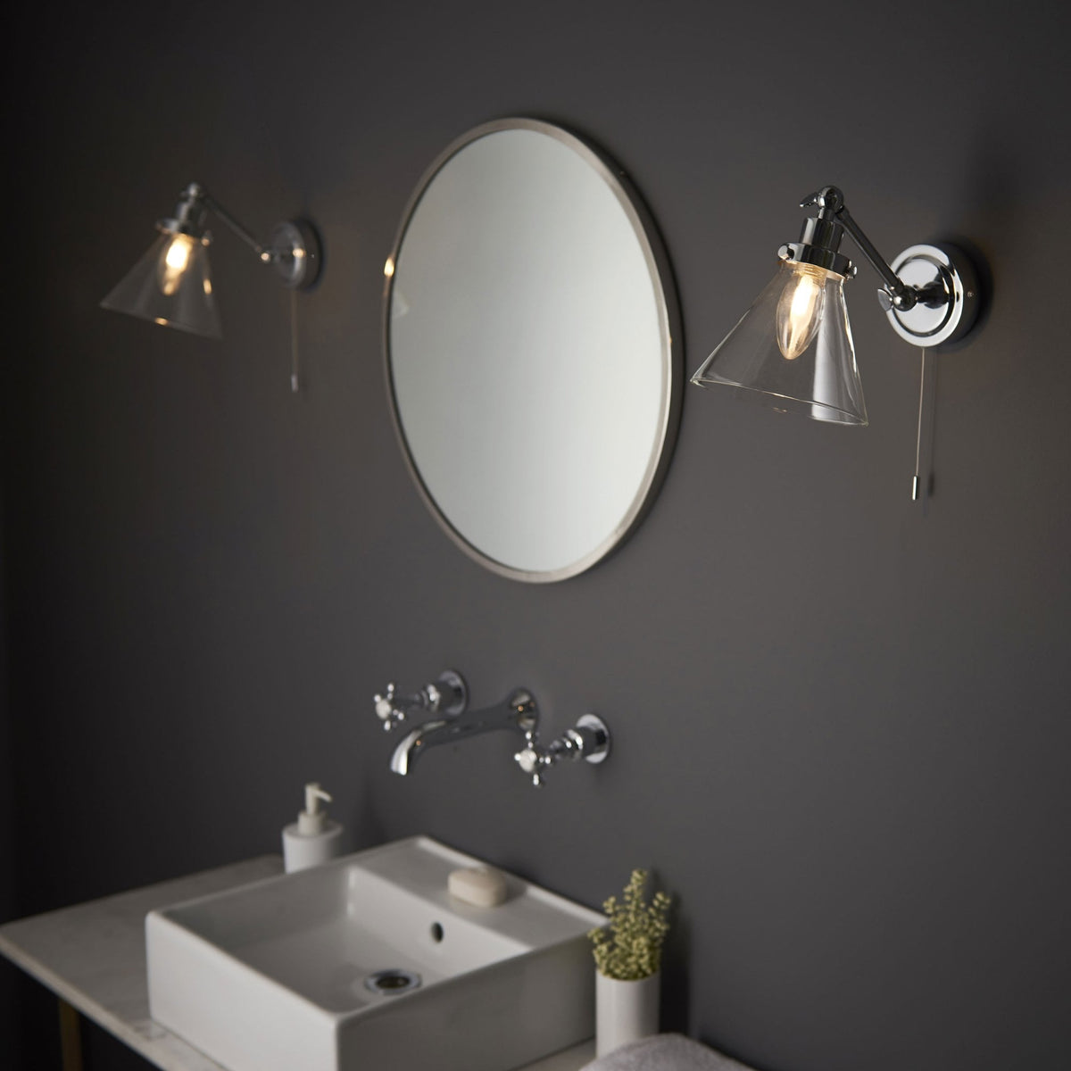 Two chrome wall lights with clear glass cone shades flank a round mirror above a white sink and wall-mounted faucet.
