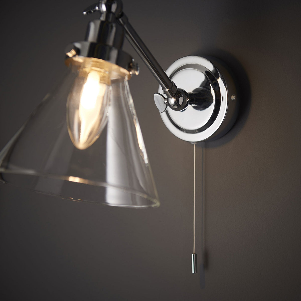 Close-up of a chrome wall light with a clear glass cone shade and pull cord switch, casting a warm glow on a dark wall.