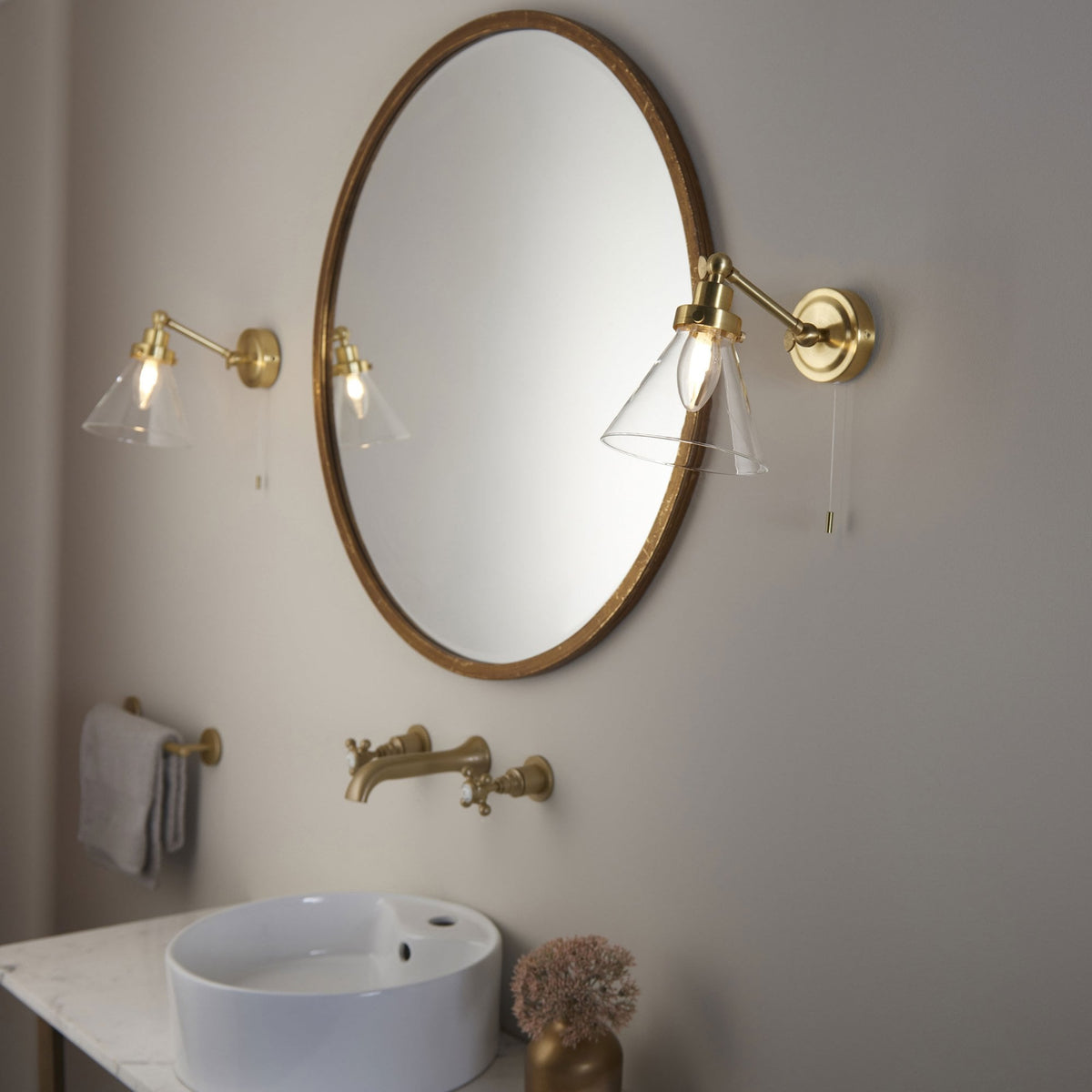 Two brass wall lights with clear glass shades flanking an oval mirror above a white sink and brass faucet.