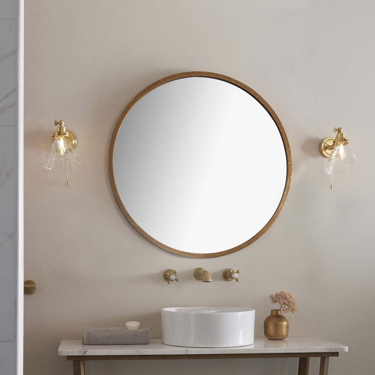 Bathroom vanity with a round mirror, two brass wall lights, a white basin, and brass wall-mounted taps.