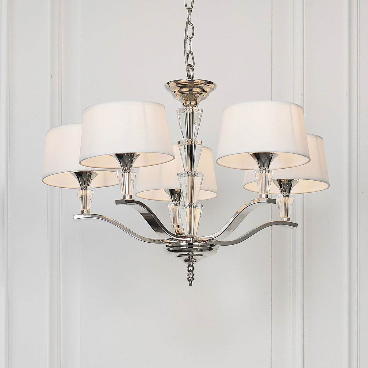 A five-arm chandelier with white fabric shades and clear crystal accents, hanging against a white wall.