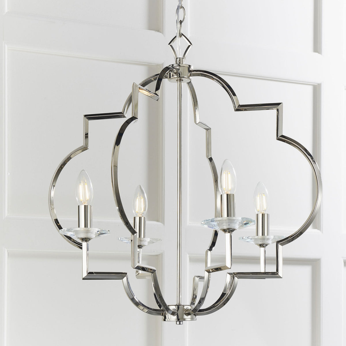Close-up of a polished nickel chandelier with four lights, geometric frame, and clear glass accents, against a white paneled wall.