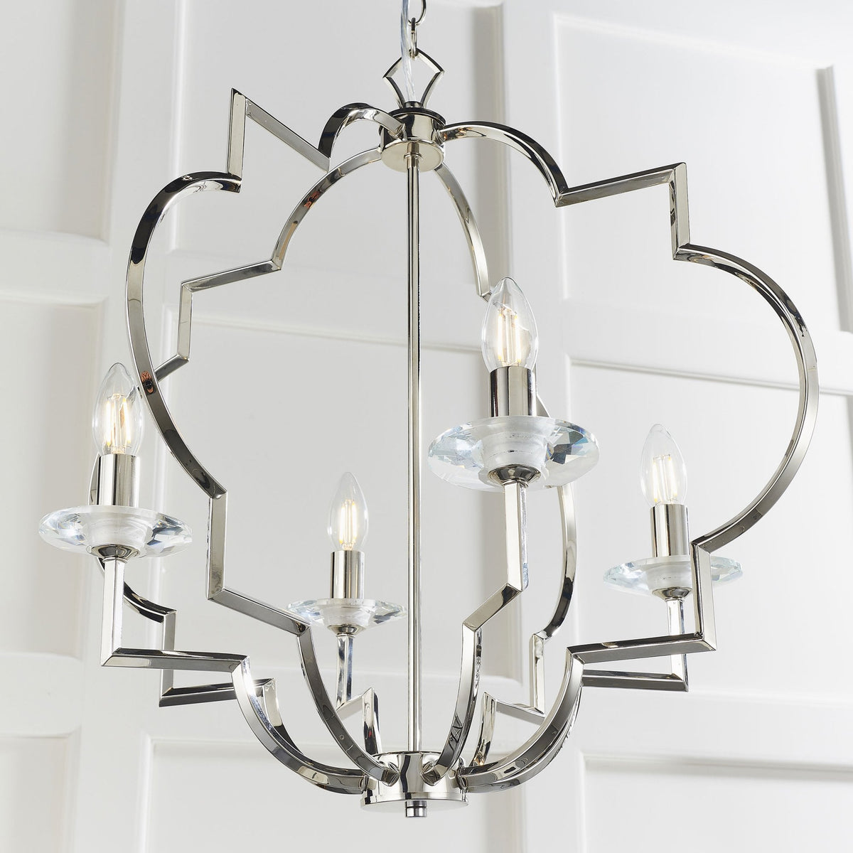 Polished nickel chandelier with four lights, geometric frame, and clear glass accents, hanging in a room with white paneled walls.