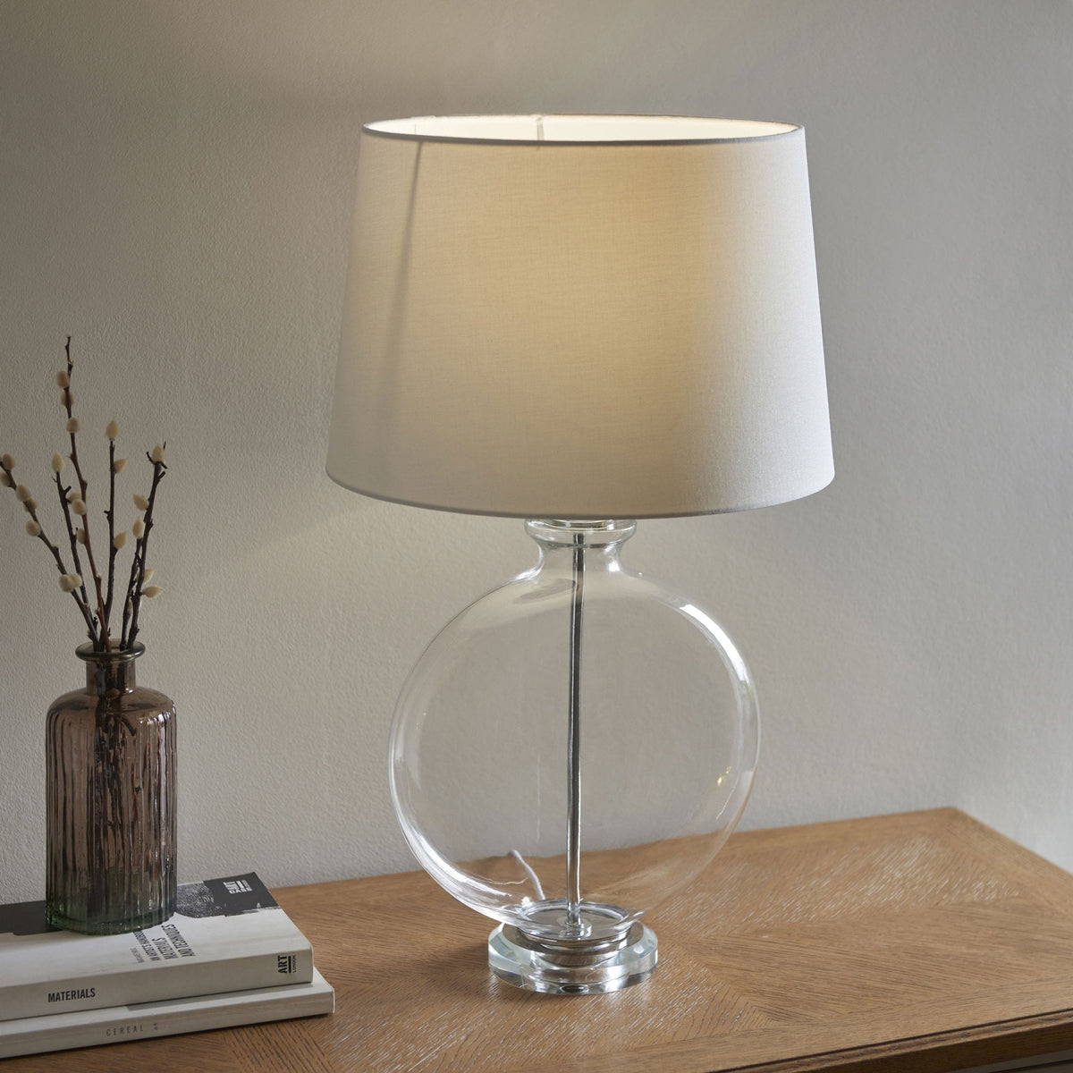 Clear glass table lamp with a round base and a white fabric shade, casting a warm glow on a wooden surface.