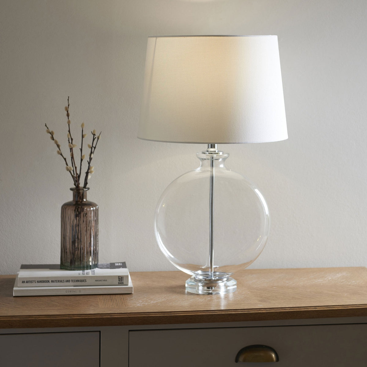 Clear glass table lamp with a round base and a white fabric shade, next to a vase with twigs on a wooden table.