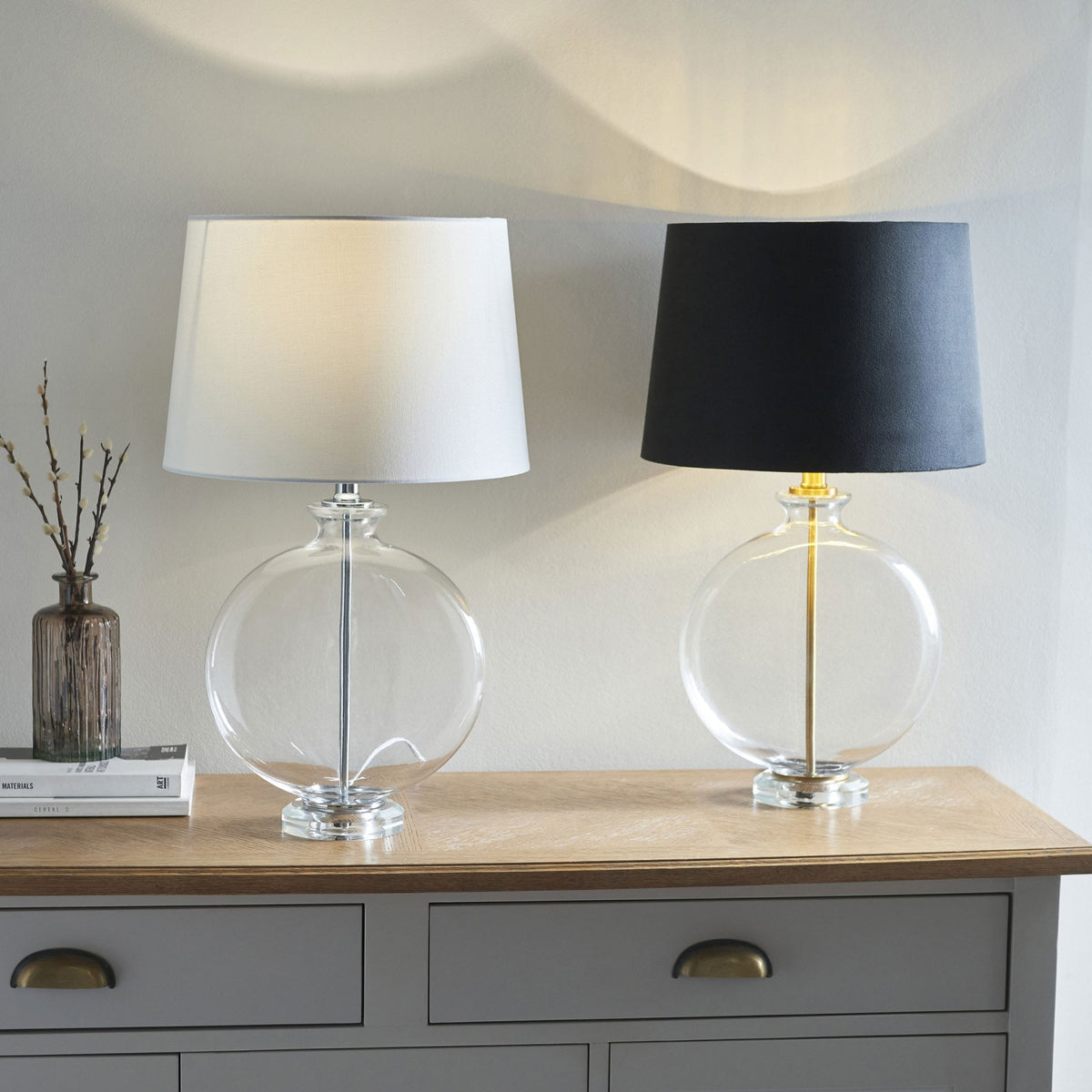 Two clear glass table lamps with round bases, one with a white shade and one with a black shade, on a wooden table.