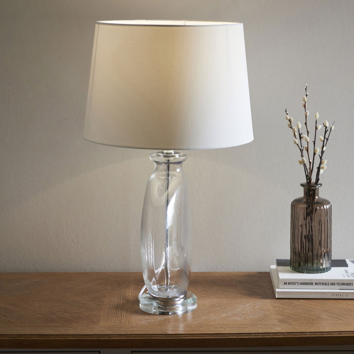 Clear glass table lamp with a twisted base and a white fabric shade, next to a vase with twigs on a wooden table.