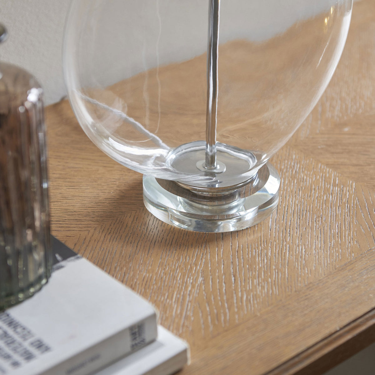 Close-up of the clear glass base of a table lamp, showing its round shape and metallic stem on a wooden surface.