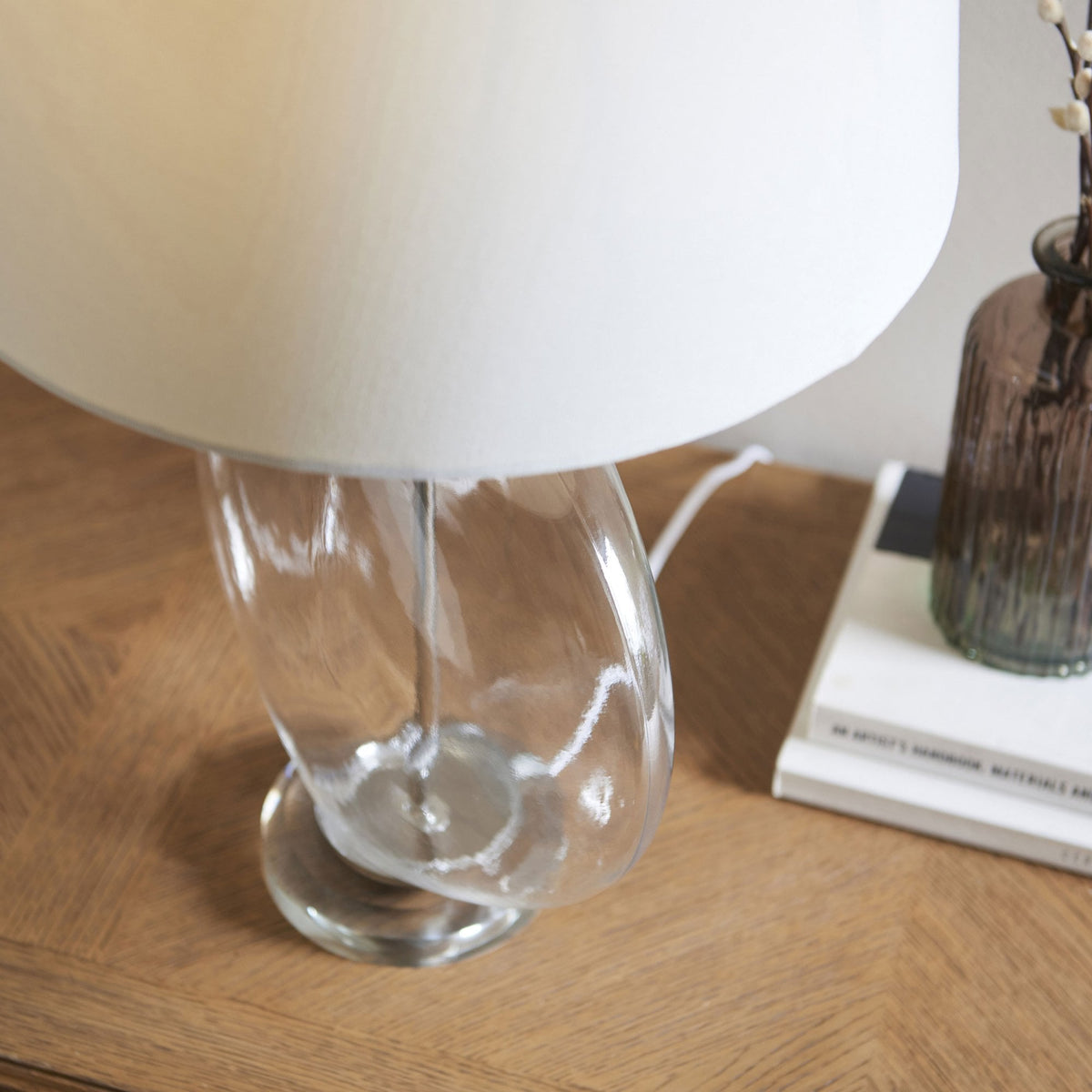 Close-up of a clear glass table lamp with a twisted base and a white shade, on a wooden table with books.