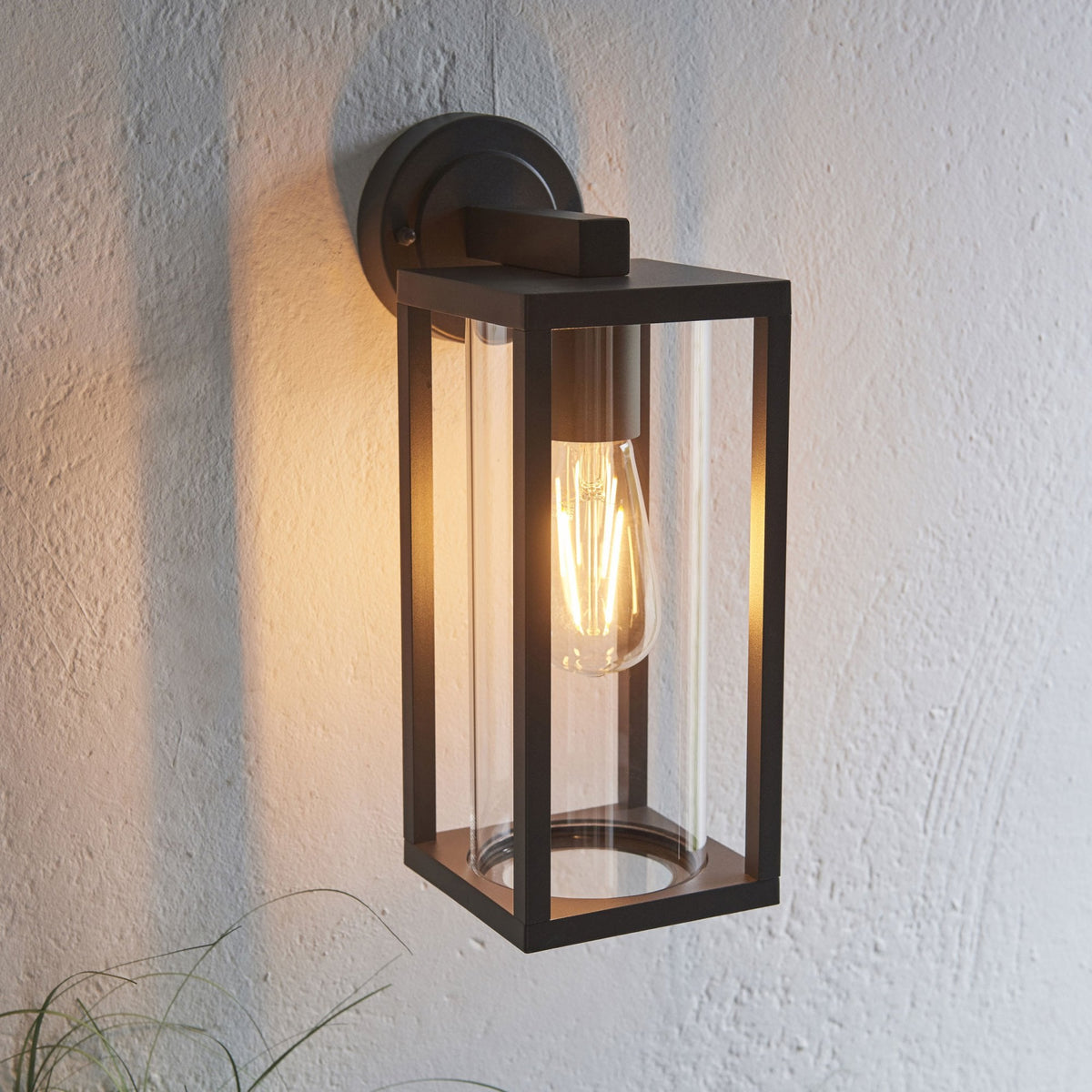 Illuminated black outdoor wall light with clear glass panels, casting a warm glow on a textured wall.