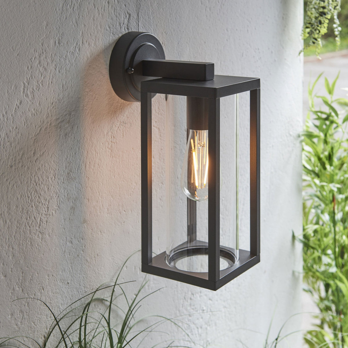 Black outdoor wall light with clear glass panels and a glowing filament bulb, mounted on a light wall.