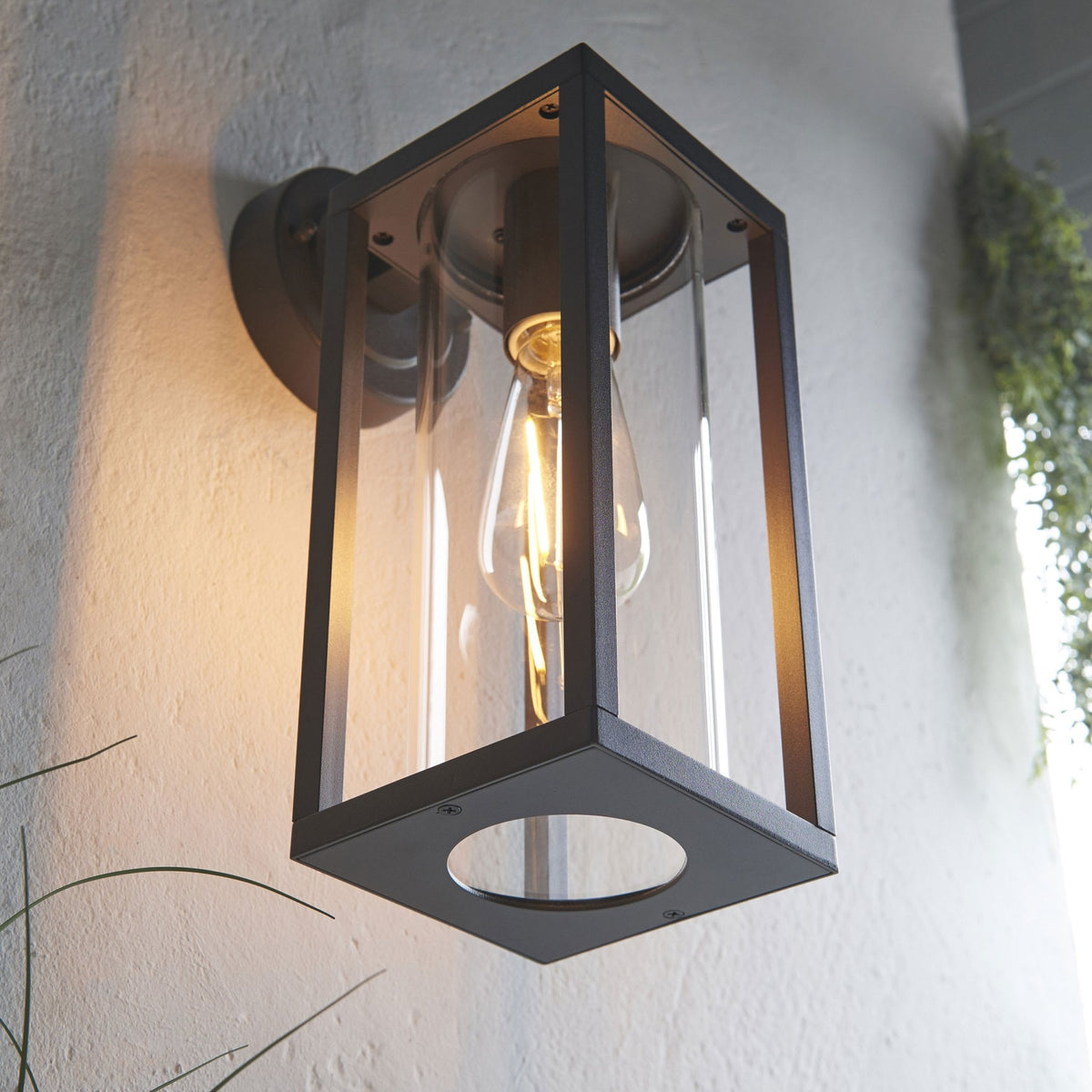 Close-up of an illuminated black outdoor wall light with clear glass panels and a visible filament bulb.