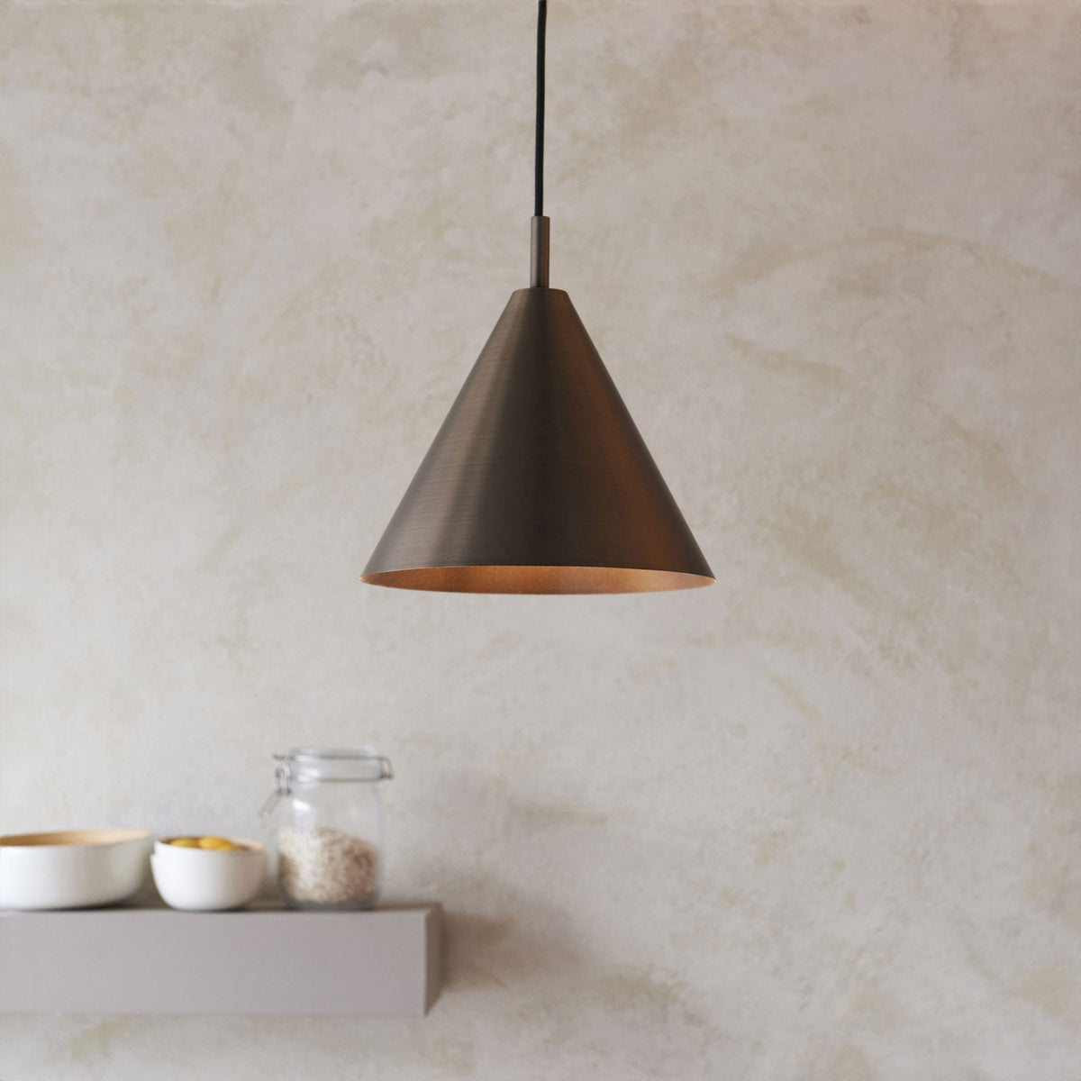 A dark bronze conical pendant light hangs above a kitchen shelf with bowls and a jar.