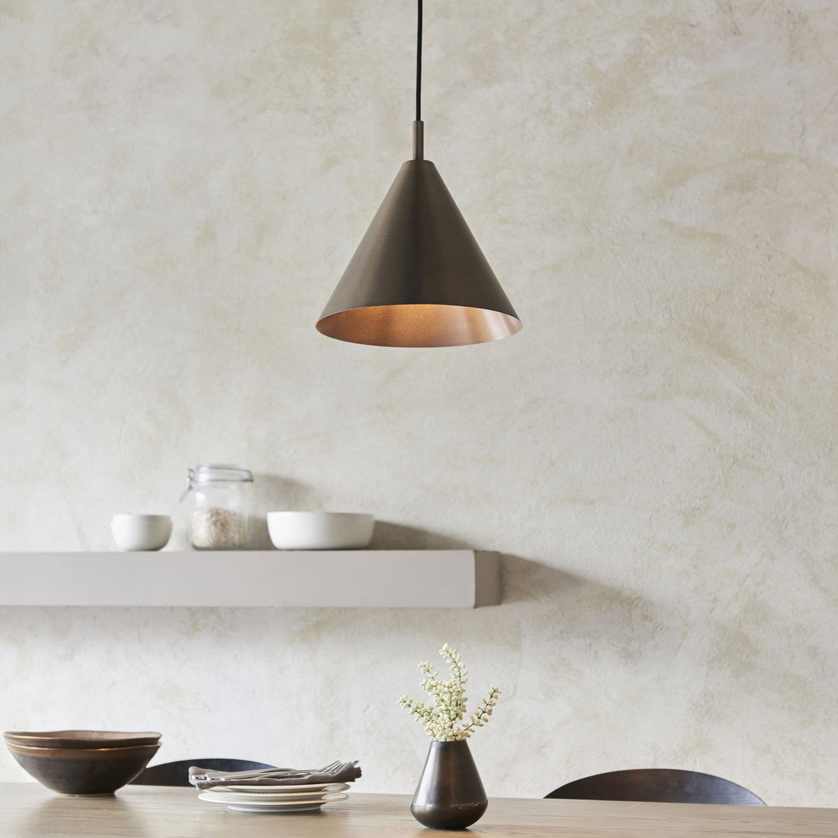 A dark bronze conical pendant light illuminates a dining table with bowls, plates, and a small vase.
