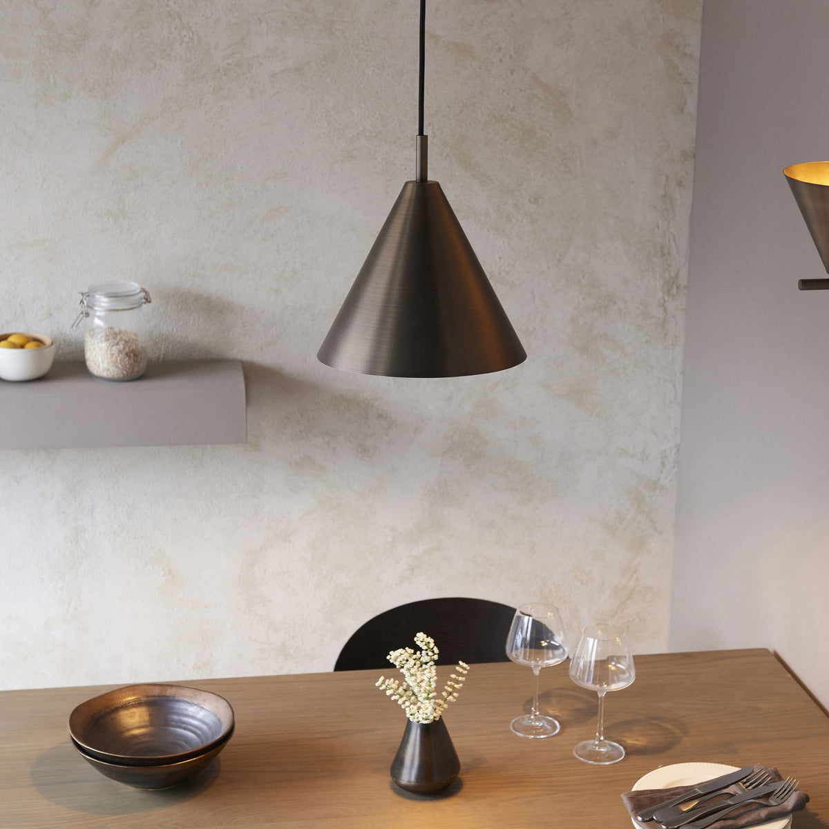 A dark bronze conical pendant light hangs above a dining table with glasses and a vase.