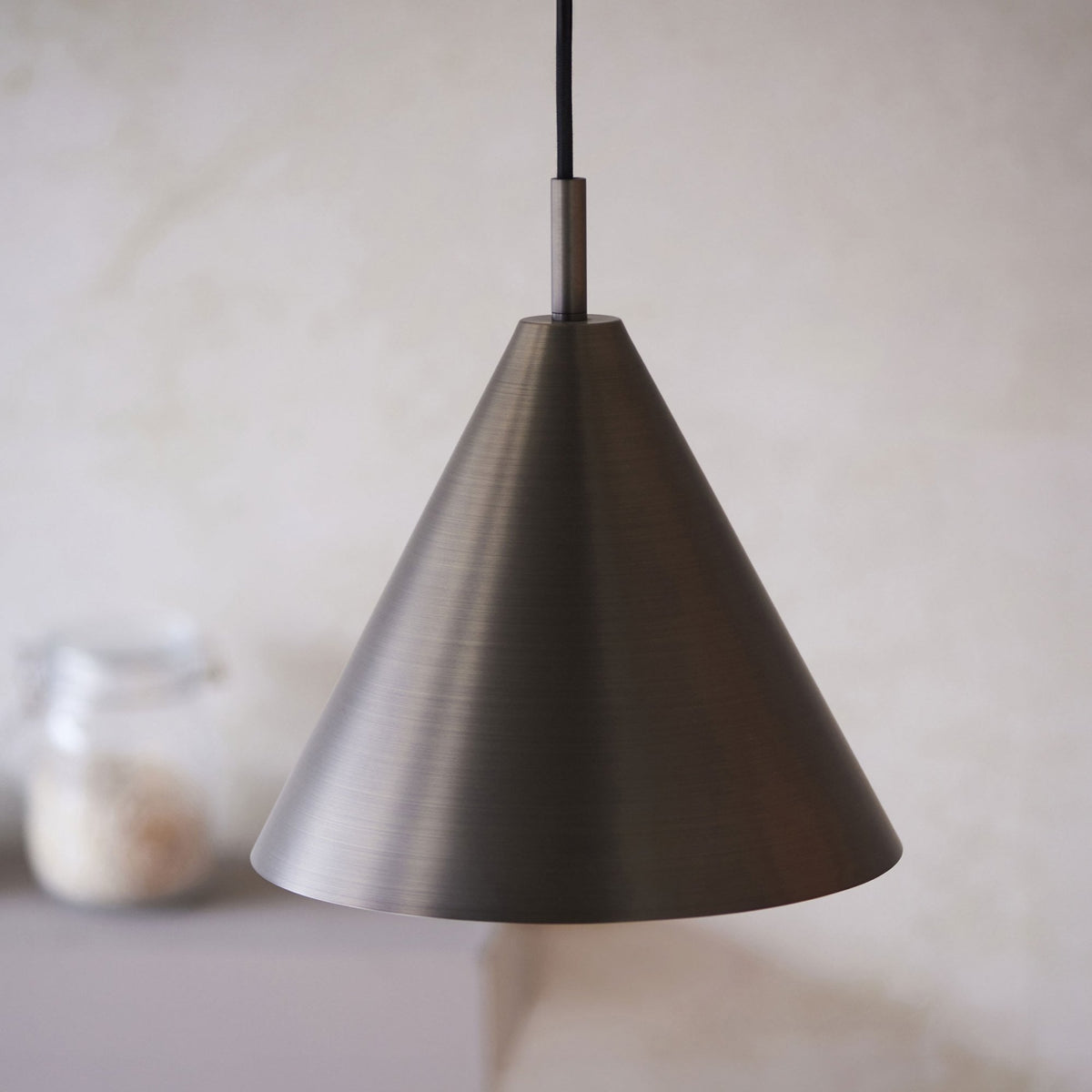 Close-up of a dark bronze conical pendant light with a brushed finish, above a blurred shelf.