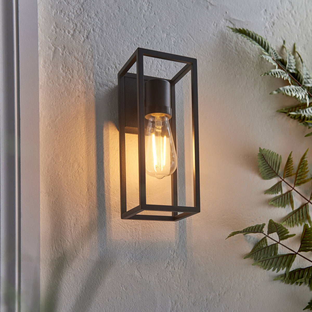 Black framed wall light with an exposed bulb illuminating a textured wall and green foliage.