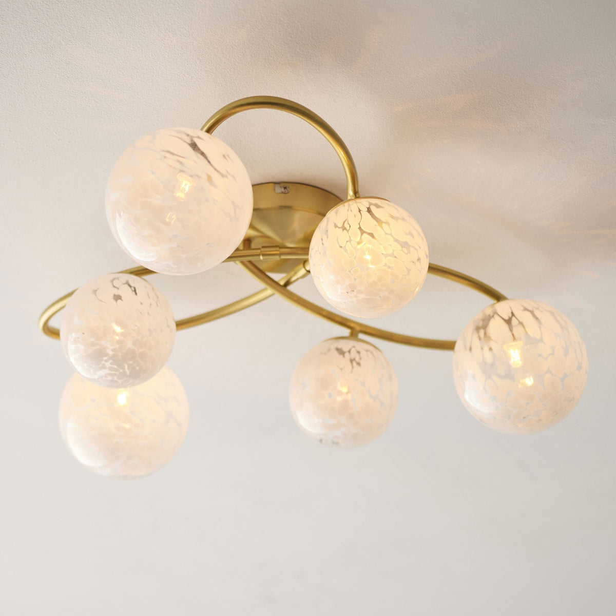 Close-up of a gold ceiling light with six white speckled glass globes, illuminated and casting a warm glow.