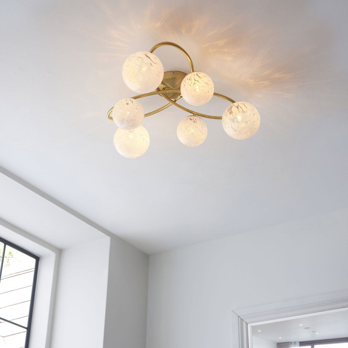 Gold ceiling light with six white speckled glass globes, illuminated, in a bright room with a window.