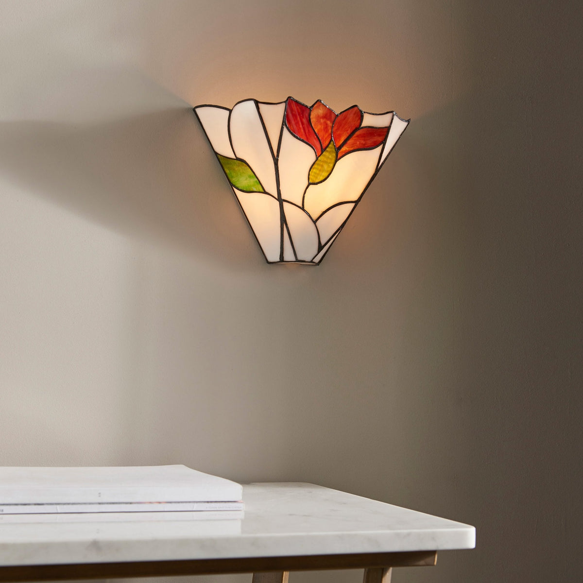 A stained glass wall light with a red flower and green leaves mounted on a wall above a table.