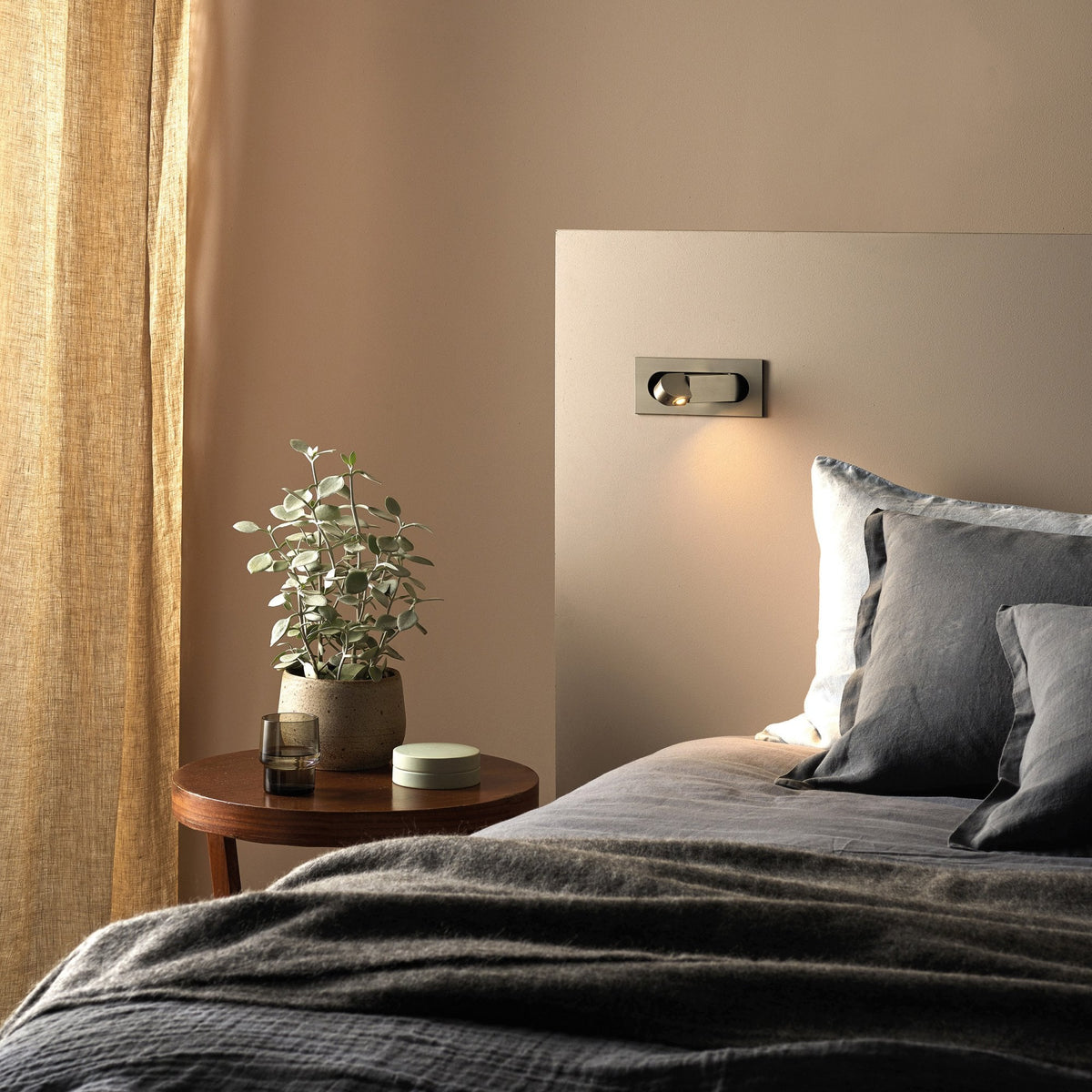 Bedroom with beige walls, a bed with grey bedding, a wooden side table, and a plant