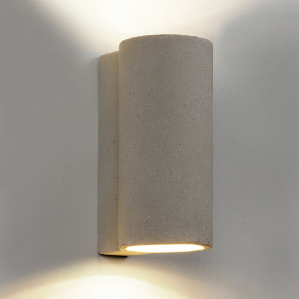A modern cylindrical concrete up-and-down wall light casting warm light on a white wall.