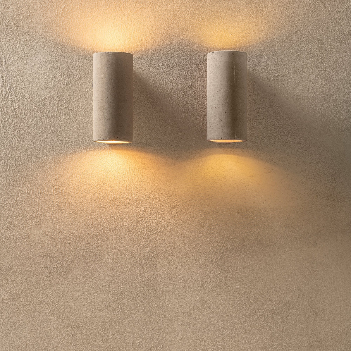 Two identical cylindrical concrete up-and-down wall lights illuminating a textured wall.