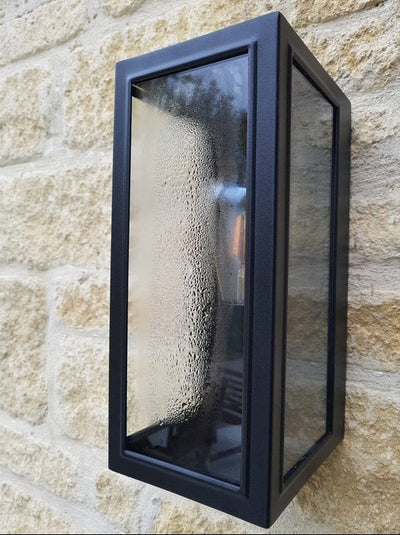 Black rectangular outdoor wall light with clear glass panels mounted on a stone wall