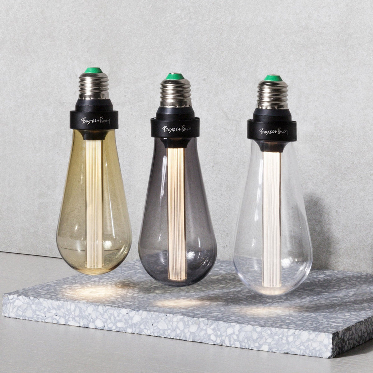 Three Buster + Punch LED light bulbs in amber, smoked, and clear glass, casting light on a terrazzo surface.
