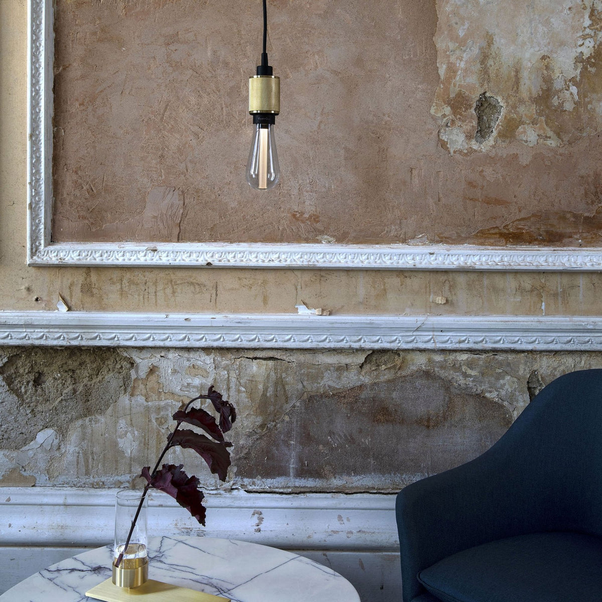 A brass pendant light with an exposed bulb hangs above a marble table with a dark leaf in a vase. 