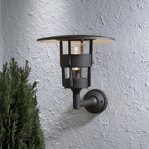 A black outdoor wall light with an open design and a subtle glow, next to a green bush.