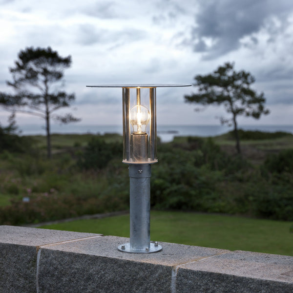 A bollard lamp illuminates a stone wall by the sea at dusk.