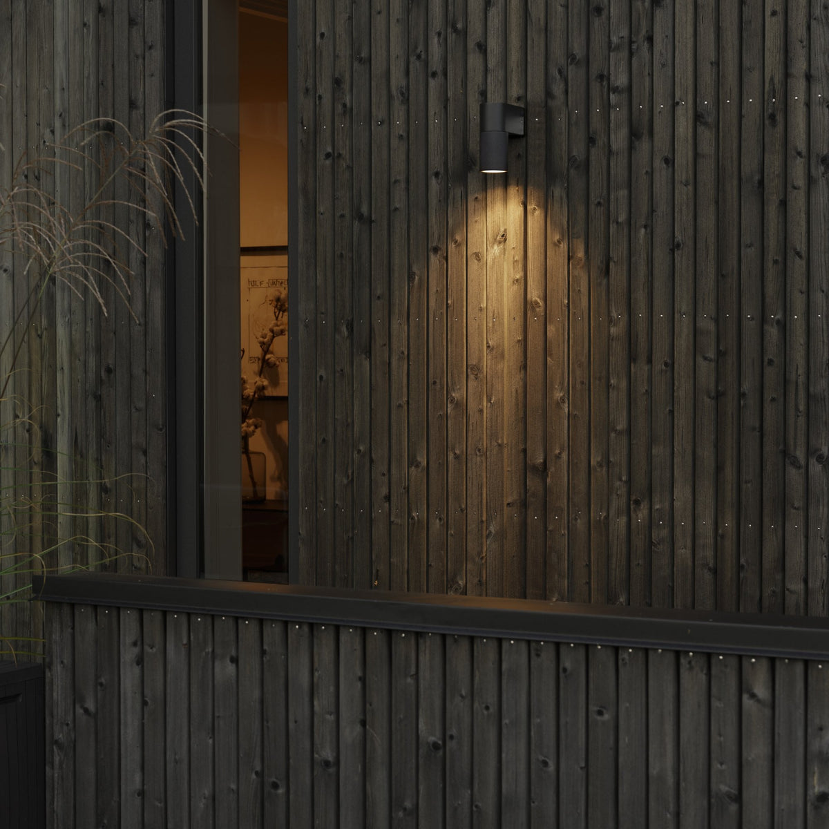 A black wall light casting a warm glow on a dark wooden-slatted exterior wall at dusk.