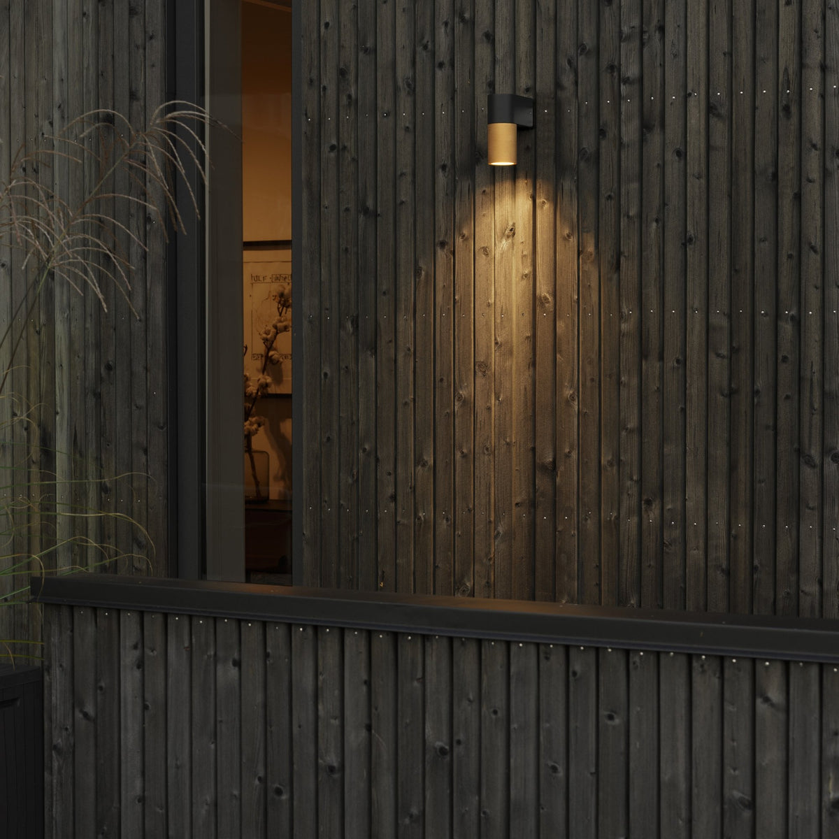A gold and black wall light illuminating a wooden slatted exterior wall at dusk.