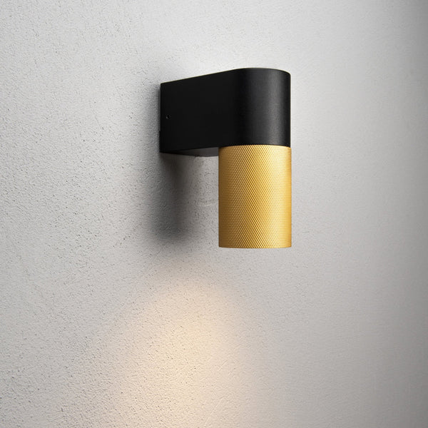 A black and gold wall light illuminating a white wall, showcasing its warm glow.